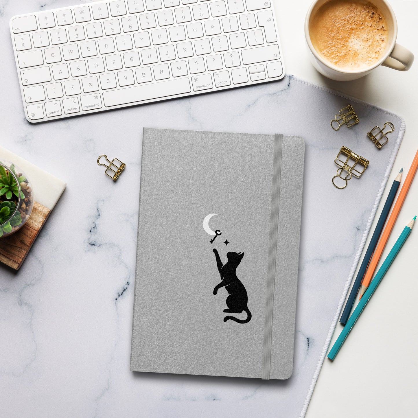 Curious Things hardcover notebook in silver with a black cat and moon illustration, styled on a soft blanket with a pen, instant photos, and earbuds.