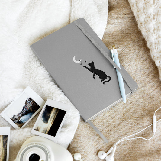Curious Things hardcover notebook in silver with a black cat and moon illustration, styled on a soft blanket with a pen, instant photos, and earbuds.