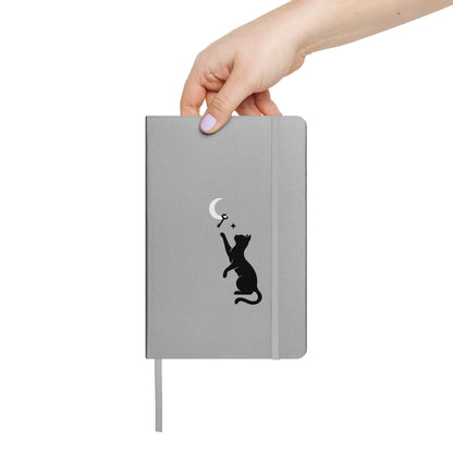 Hand holding the Curious Things silver hardcover notebook featuring a black cat reaching toward a crescent moon.